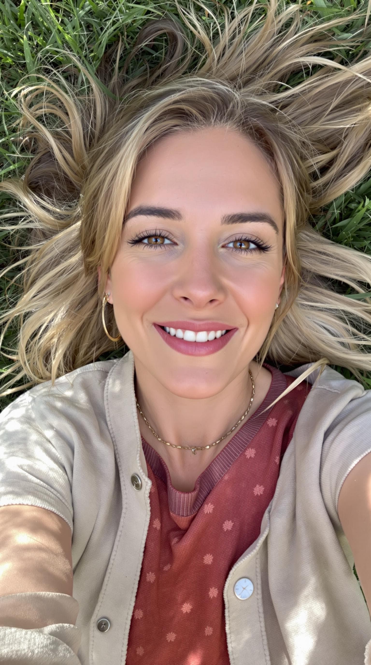 A point-of-view image as if taken from a smartphone, showing a woman in her mid-30s talking directly to the camera in a selfie-style pose, outside shot. She has a warm and expressive face with a slight head tilt and a confident smile, as if recording a vlog or leaving a heartfelt message. The background laying on grass